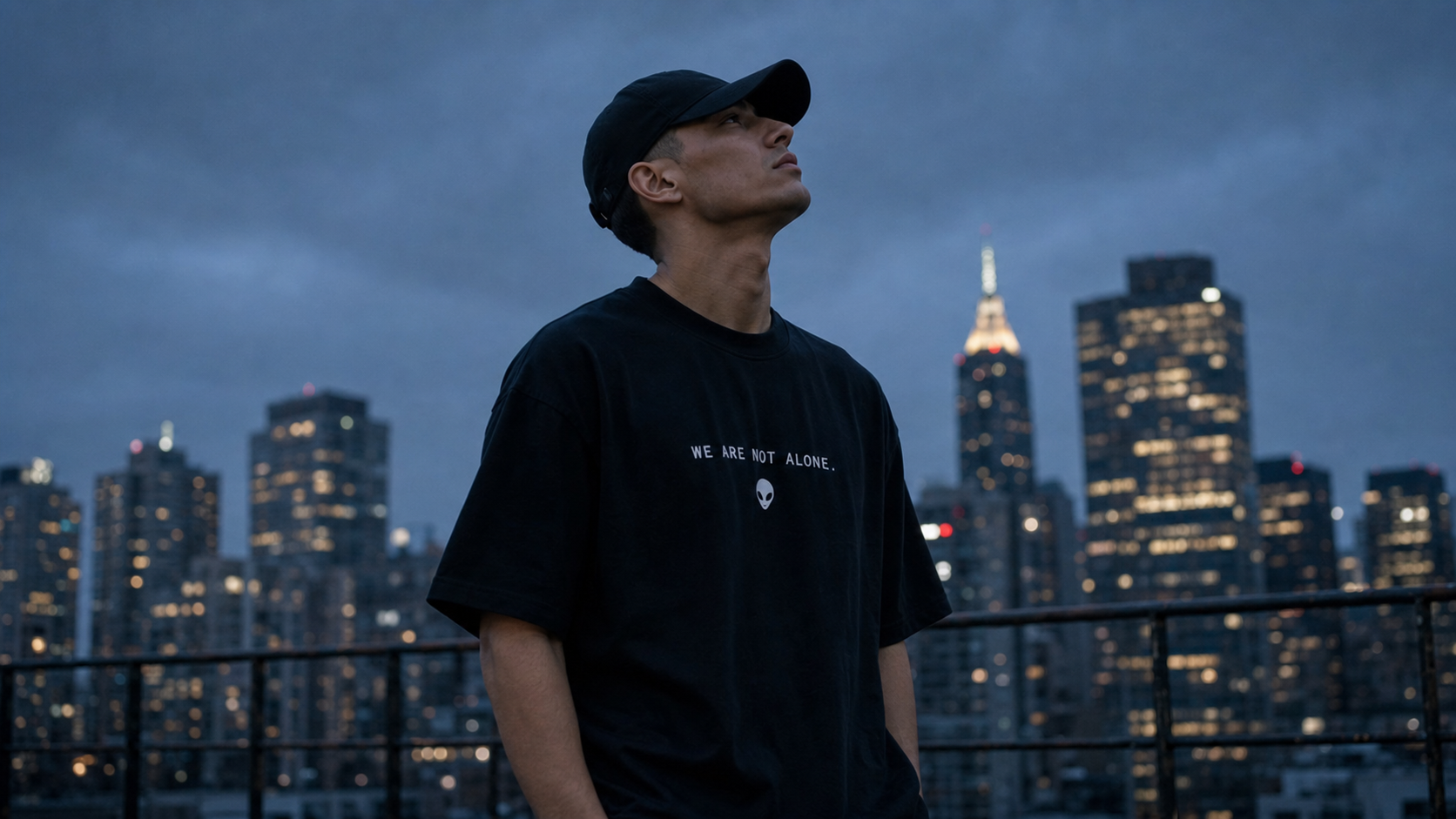 First Contact Club model wearing the black We Are Not Alone T-shirt on a rooftop at blue hour