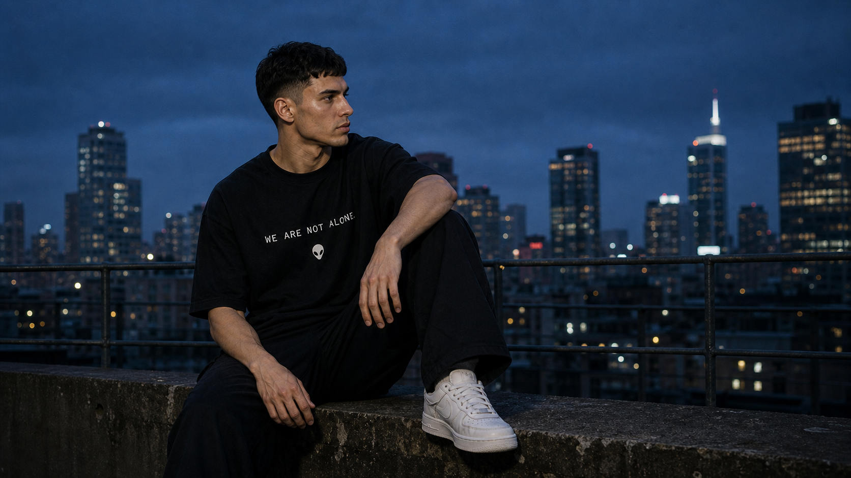 First Contact Club model seated naturally on a rooftop ledge wearing the black We Are Not Alone T-shirt